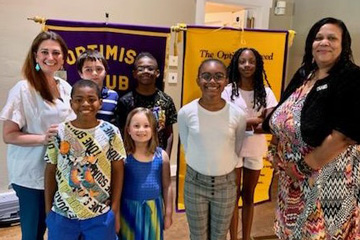 Principal and teacher standing with elementary students in front of Optimist Club banner