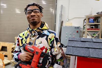 Smiling student holding nail gun in front of Little Free Library construction project