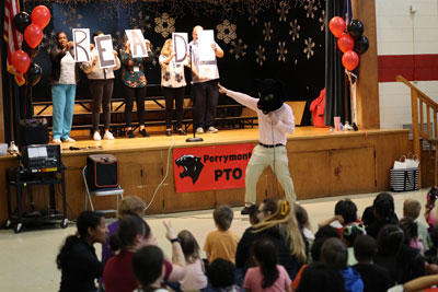 School administrator wearing school mascot costume pointing to people on stage holding signs spelling out "READ"