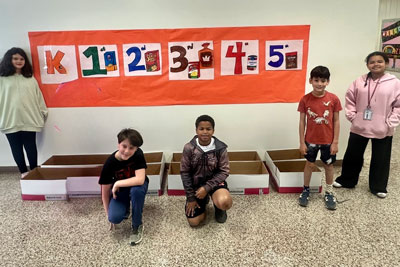 Four students standing around boxes and banner labeled K, 1, 2, 3, 4, 5