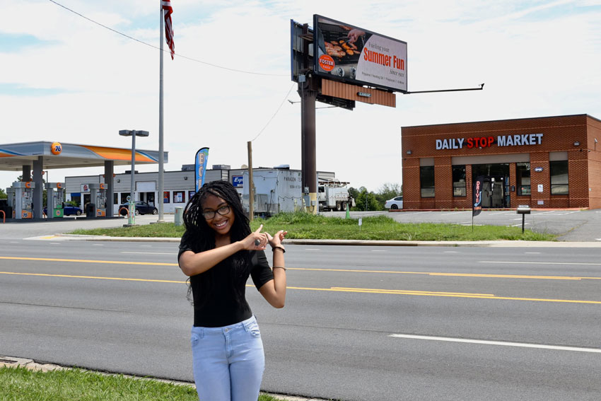 Student standing outside pointing at billboard in background