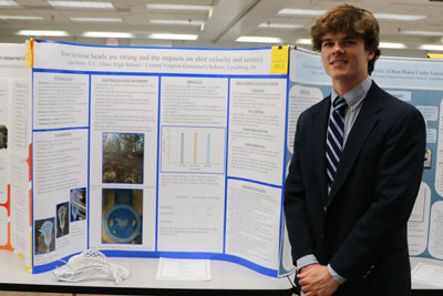 High school student in professional attire standing in front of science fair display with title “How Lacrosse Heads are Strung and the Impacts on Shot Velocity and Accuracy”