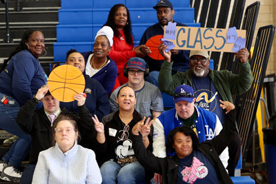 Group of adults sitting on bleachers, some holding basketball and "Glass" signs