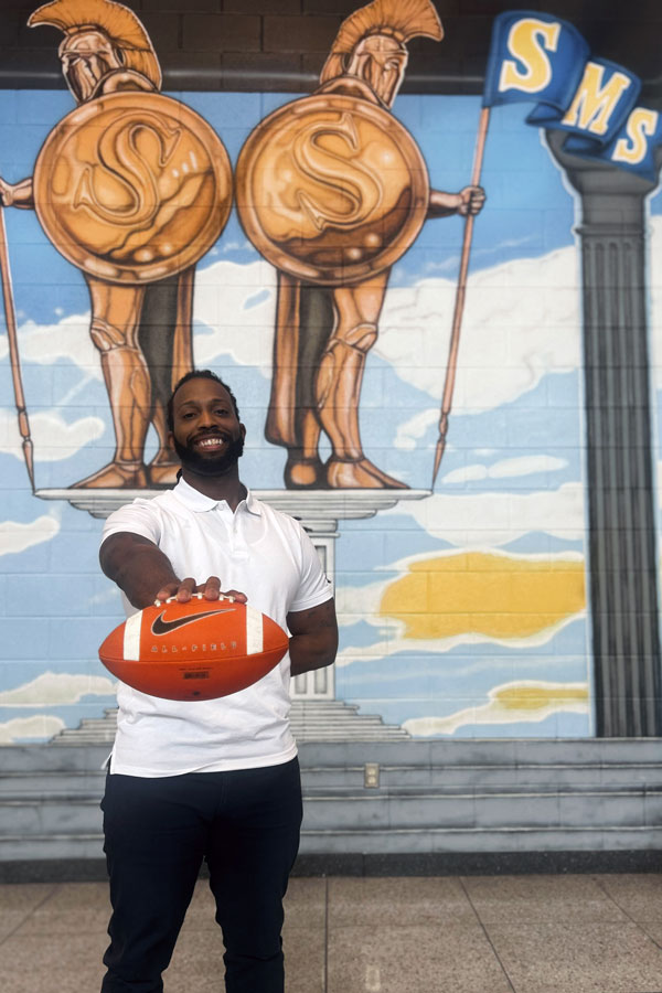 Coach Kenneth “KJ” Bryant holding football in front of Sandusky Middle School mural