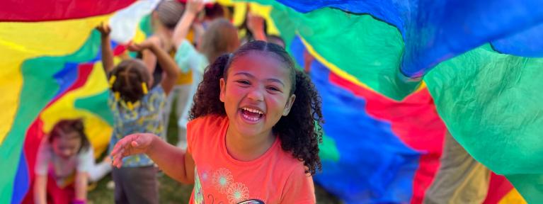 Smiling girl with other students playing under colorful parachute