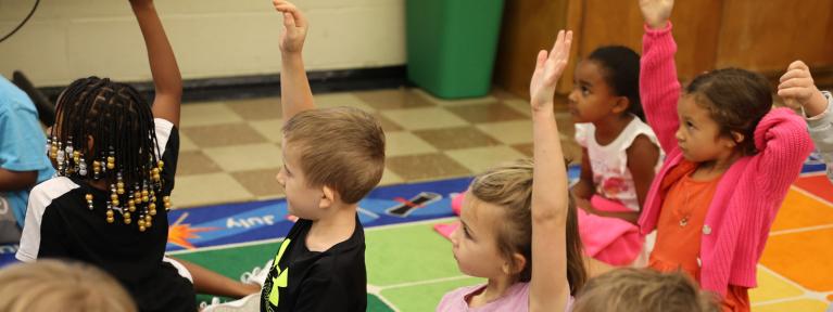 Students sitting on colorful rug raising hands