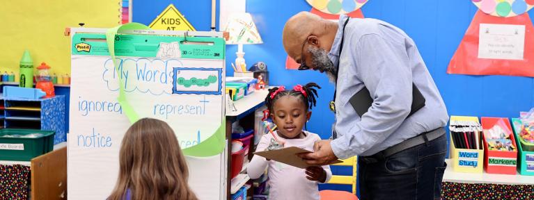 Principal in colorful classroom holding a clipboard on which a student is writing